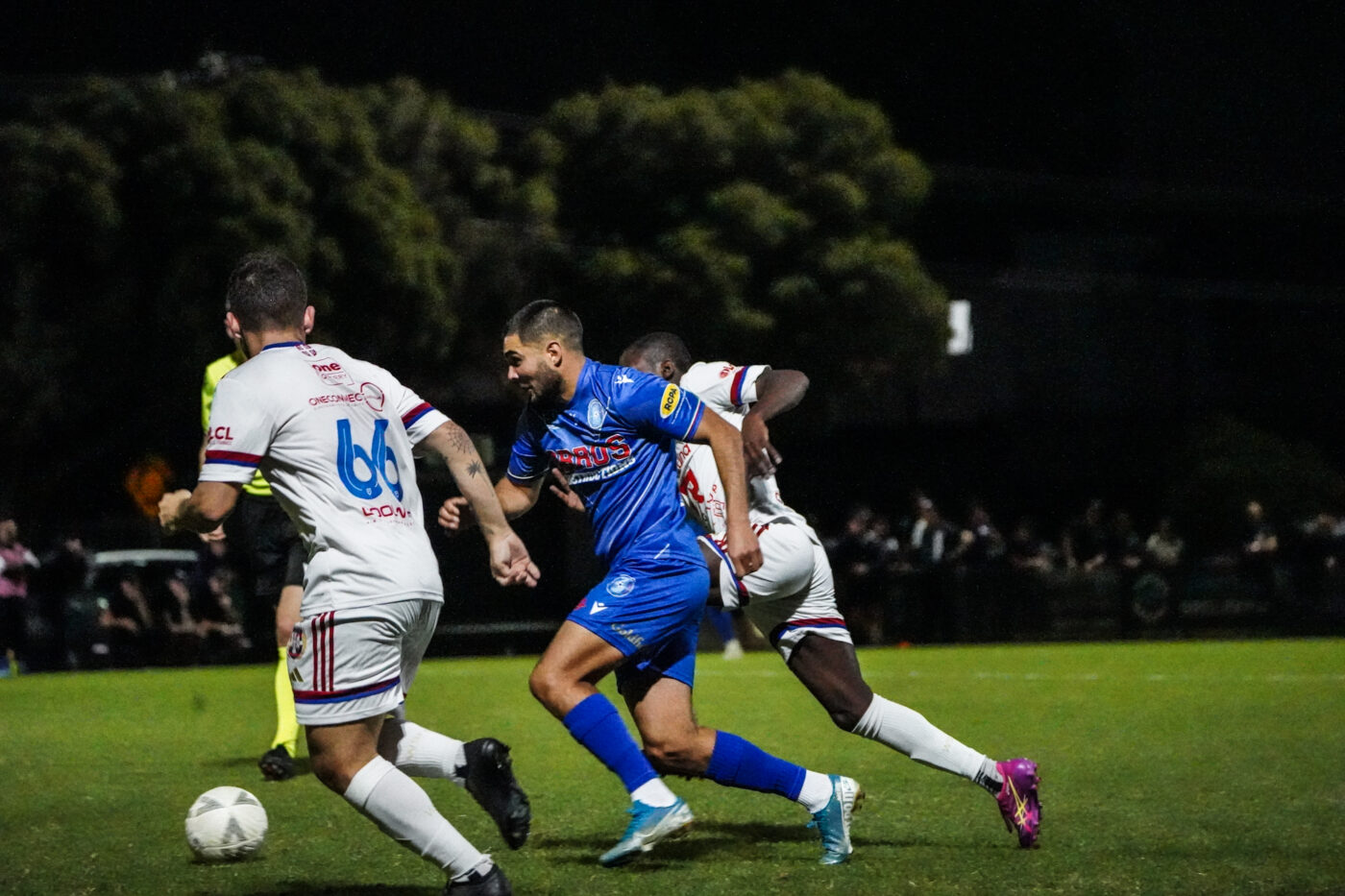 Avondale FC player in blue kit dribbles past opponents under the lights during a night match.