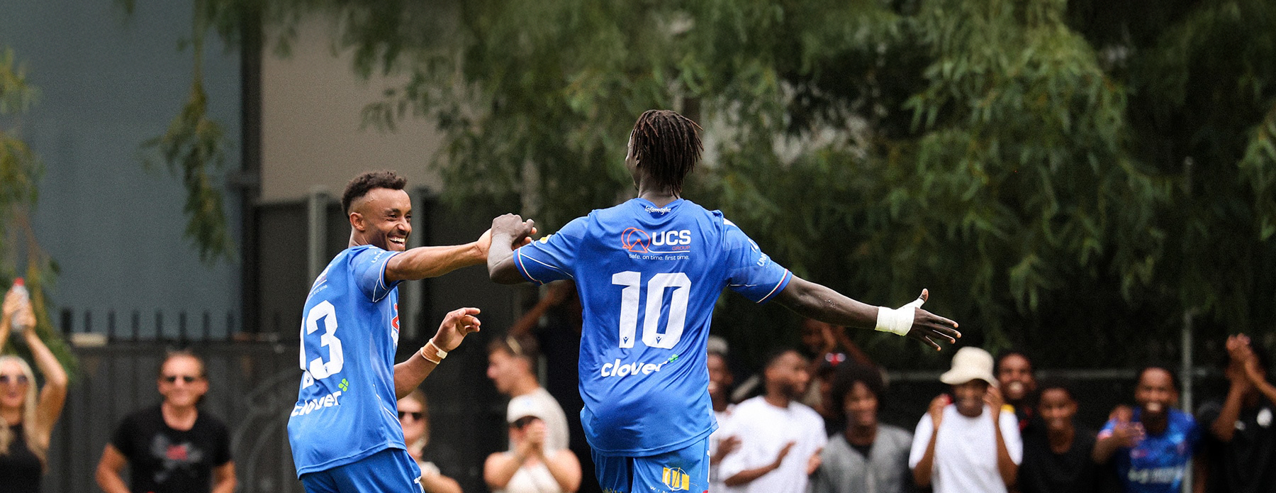 Joyful Avondale FC teammates embrace after scoring as supporters applaud from the stands.