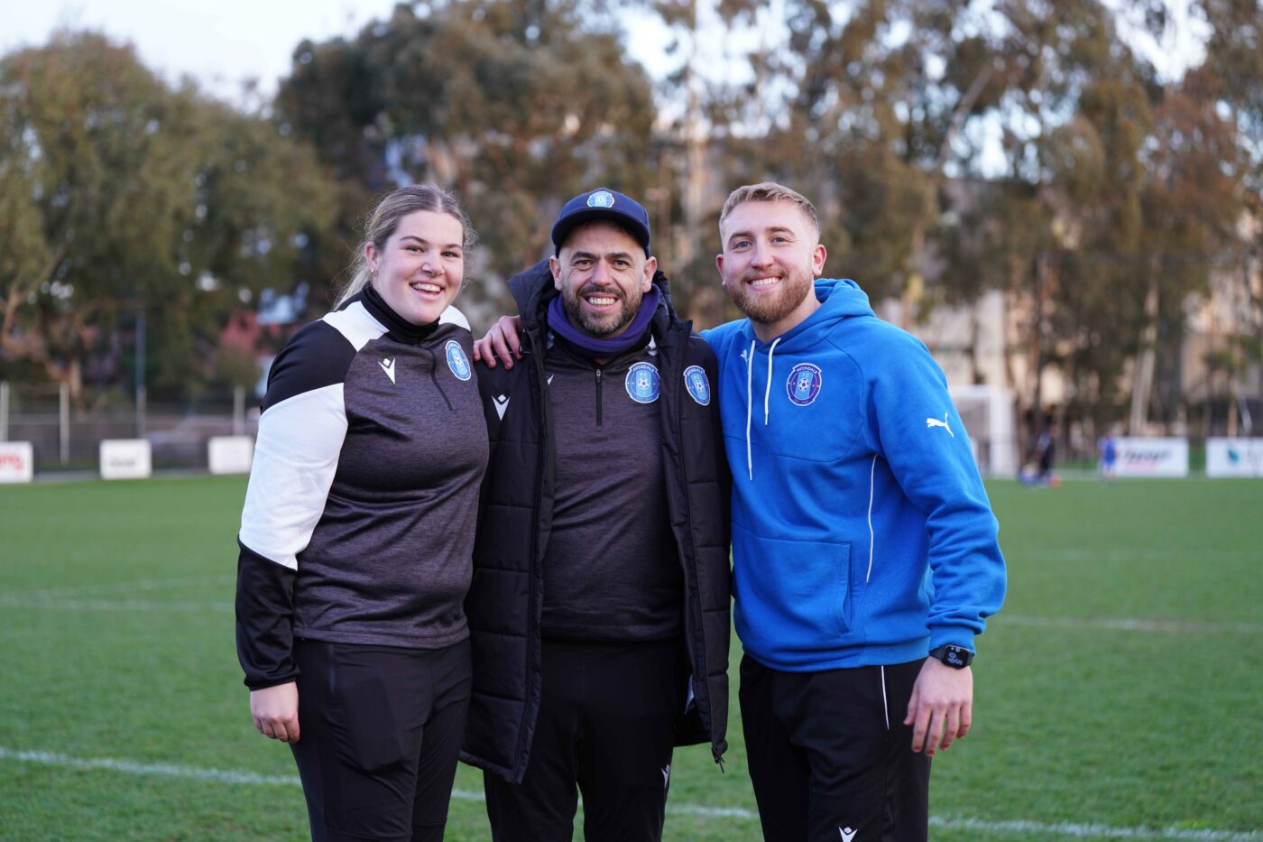 Three Avondale FC coaches standing arm in arm on the pitch, happy and supportive.